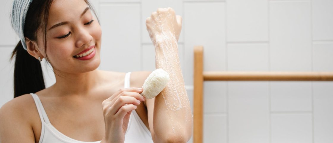 Imagem de uma mulher com traços asiáticos dentro de um banheiro mostrando como descolorir os pelos. Ela veste roupa branca e higieniza o braço aplicando um produto de cor branca em um de seus braços com um algodão.