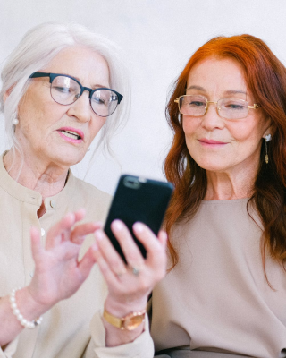 A imagem mostra três mulheres sentadas, mais maduras, cada uma com um cabelo diferente, olhando para a tela de um celular. As três usam óculos e vestem roupas de cores neutras.