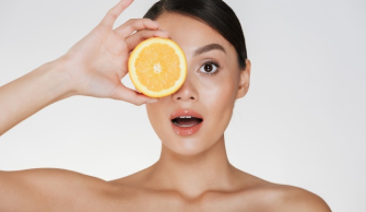 Na imagem, uma mulher segura metade de uma laranja em frente ao olho direito, diante de um fundo branco.