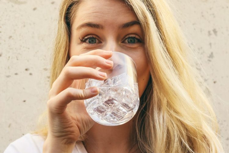 Close up de uma mulher branca, loira de cabelos compridos e olhos azuis, vestindo uma camiseta branca e tomando um copo de água.
