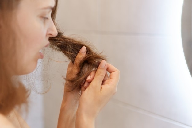 Imagem de uma mulher, vista de lado, enquanto segura com as mãos as pontas de seu cabelo e as observa descontente. Ela está localizada dentro de um banheiro em frente a um espelho.