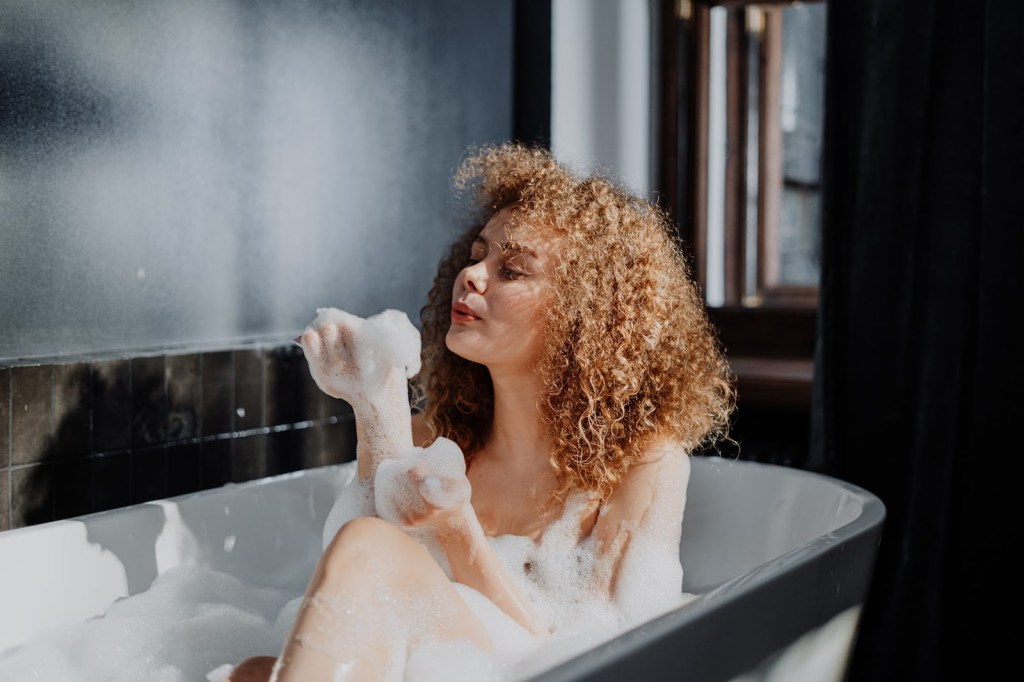 Imagem de uma mulher tomando banho de espuma em uma banheira. Ela tem cabelos cacheados e está assoprando a espuma que está em sua mão.