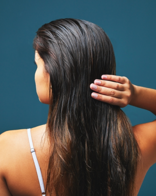 Mulher de costas tocando no cabelo oleoso pensando em como tirar oleosidade do cabelo. Ela veste sutiã branco e está contra um fundo azul.