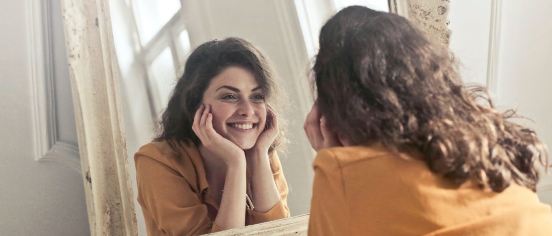Mulher sorrindo em frente a um espelho feliz por manter cuidados femininos.