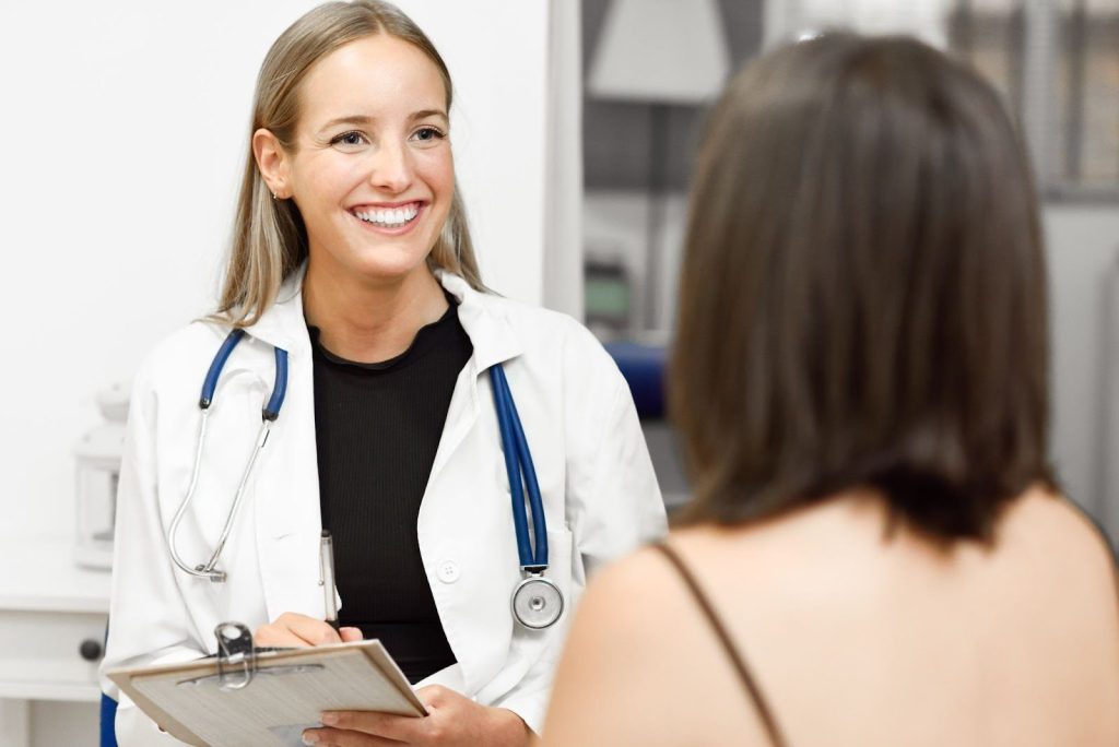 Imagem de uma mulher que mantém cuidados femininos em um consultório conversando com uma médica. A médica tem expressão feliz e faz anotações em uma prancheta.