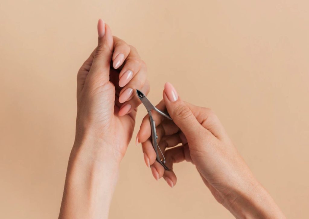 Imagem das mãos de uma pessoa branca, sobre um fundo bege. Uma das mãos segura um alicate de unhas, e aponta para as cutículas da outra mão.