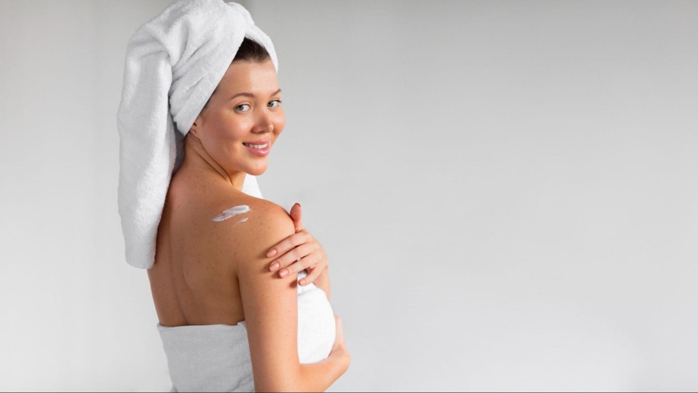 Mulher vestindo toalha de banho no corpo e no cabelo está aplicando creme hidratante no ombro como maneira de fazer o perfume durar mais.