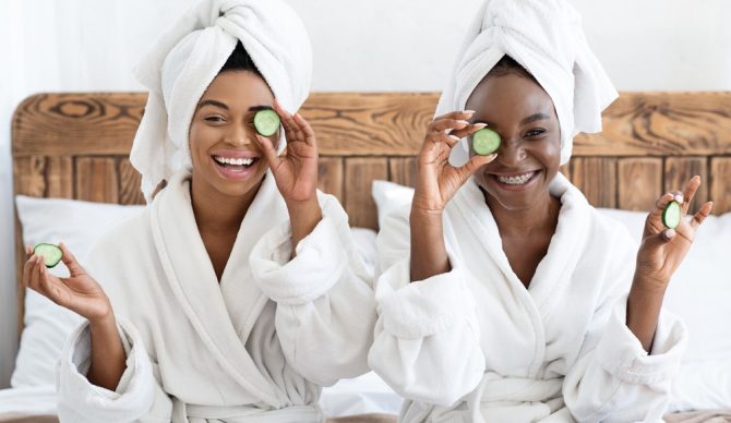 Em uma cama, duas mulheres negras estão sentadas fazendo spa em casa, usando roupões de banho e toalhas na cabeça, segurando pepinos na frente dos olhos e com máscaras faciais.