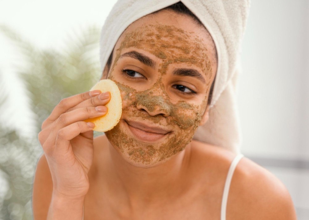 Uma mulher está fazendo spa em casa ao aplicar um esfoliante em seu rosto enquanto sorri e usa uma toalha na cabeça.