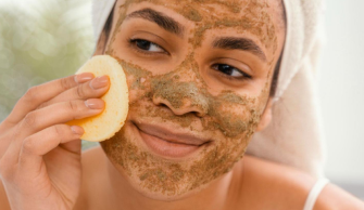 Mulher com toalha enrolada no cabelo e produto para esfoliar a pele no rosto, enquanto espalha com uma esponja.