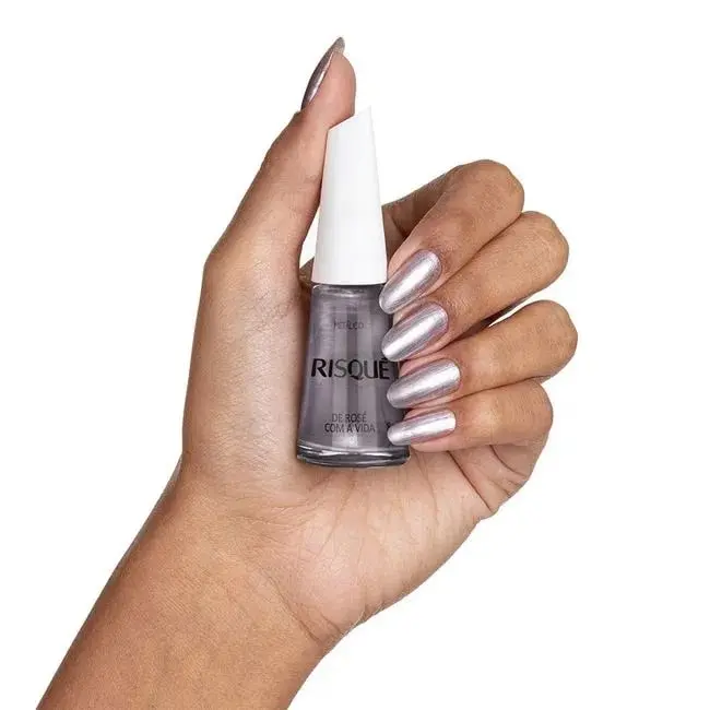 Mãos de uma pessoa branca, segurando um vidro de esmalte Risqué prata. As unhas dela estão pintadas com a mesma cor do produto.