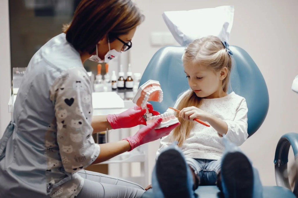 Criança está sentada em uma cadeira odontológica acompanhada por uma dentista, está segurando uma escova de dentes para entender como funciona a escovação.