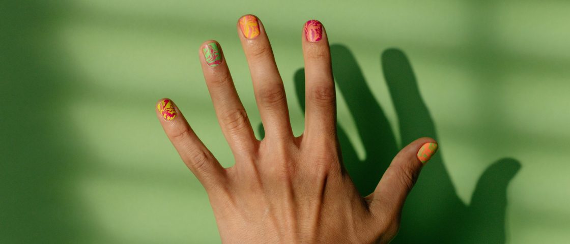 Mão de uma mulher na frente de um fundo verde e as unhas pintadas com combinações diferentes de cores de esmalte.