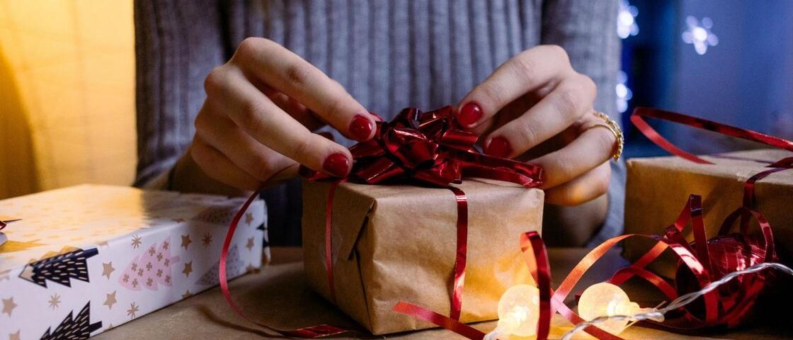 Mulher embrulhando um presente em papel pardo com laço vermelho. Há algumas luzes de Natal na mesa e outras duas caixas de presente.