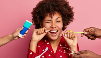 Mulher de cabelos crespos está passando o fio dental e em sua volta, 4 mãos entregam utensílios para escovar os dentes corretamente, como escova, pasta e raspador de língua.