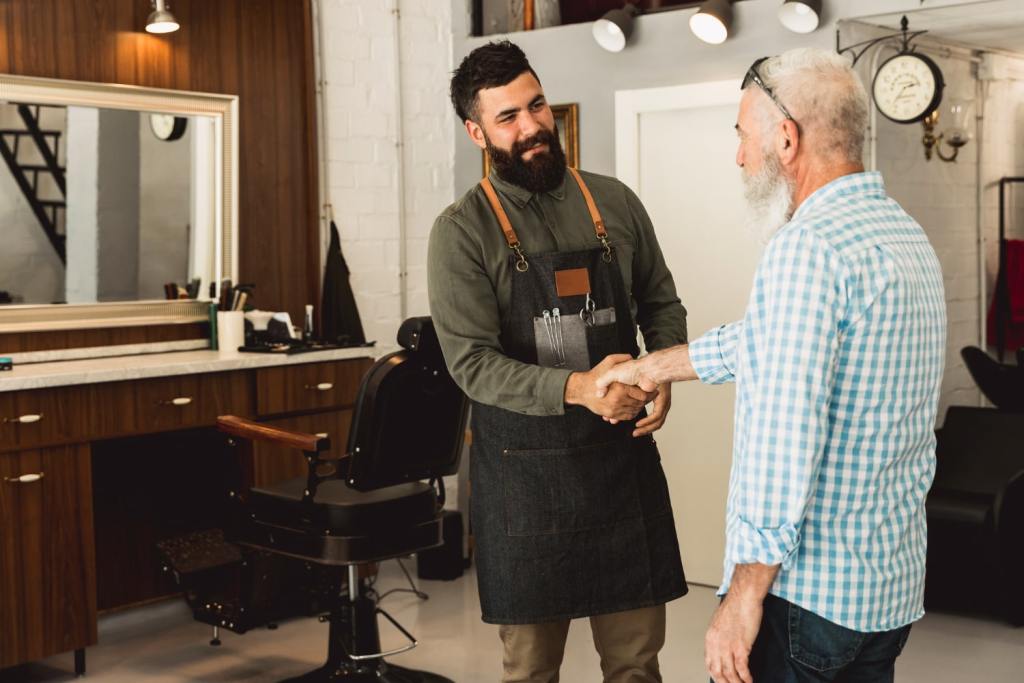 Um homem branco, já idoso, apertando a mão de seu barbeiro. Eles estão em uma barbearia, onde é possível ver a cadeira de barbeiro e o espelho ao fundo.
