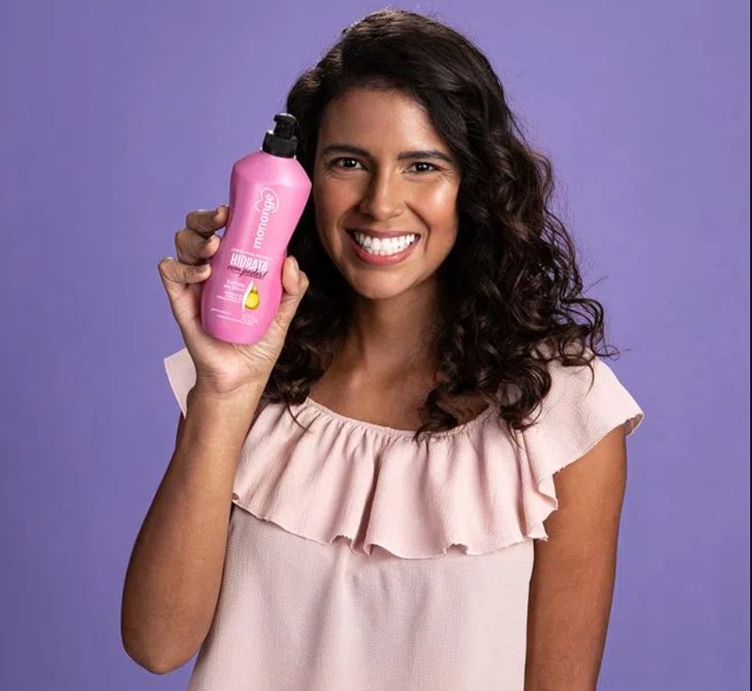 Mulher de blusa rosa, sorrindo e segurando uma embalagem do creme para pentear Monange Hidrata com poder.
