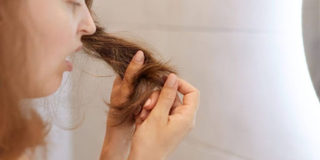 Uma mulher segurando uma mecha de seu cabelo e olhando chocada para os fios castanhos que parecem estar ressecados.