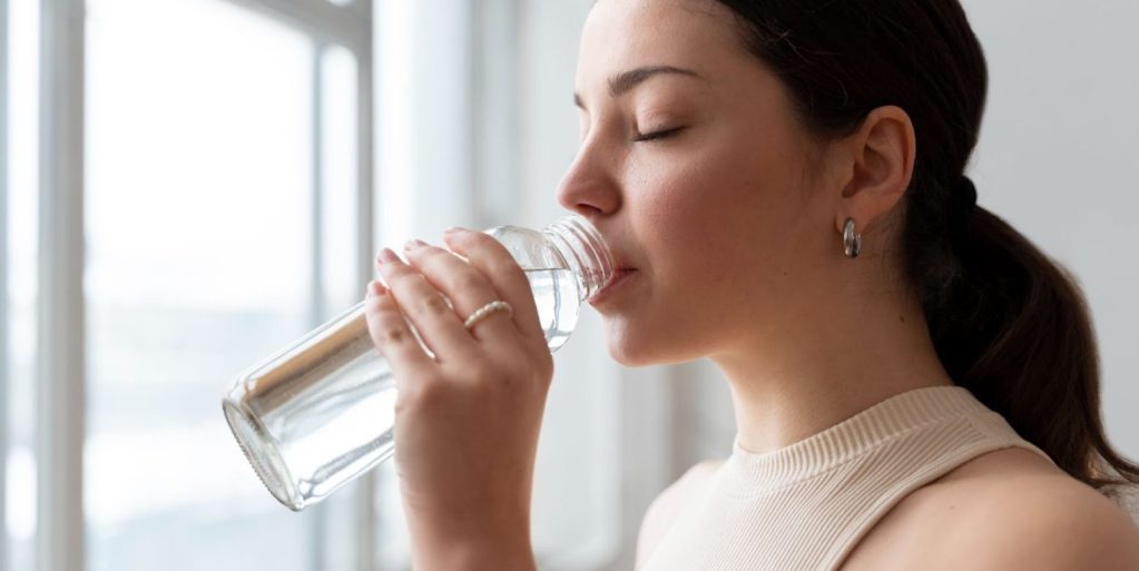 Uma mulher na frente de uma janela bebendo água em uma garrafa transparente.