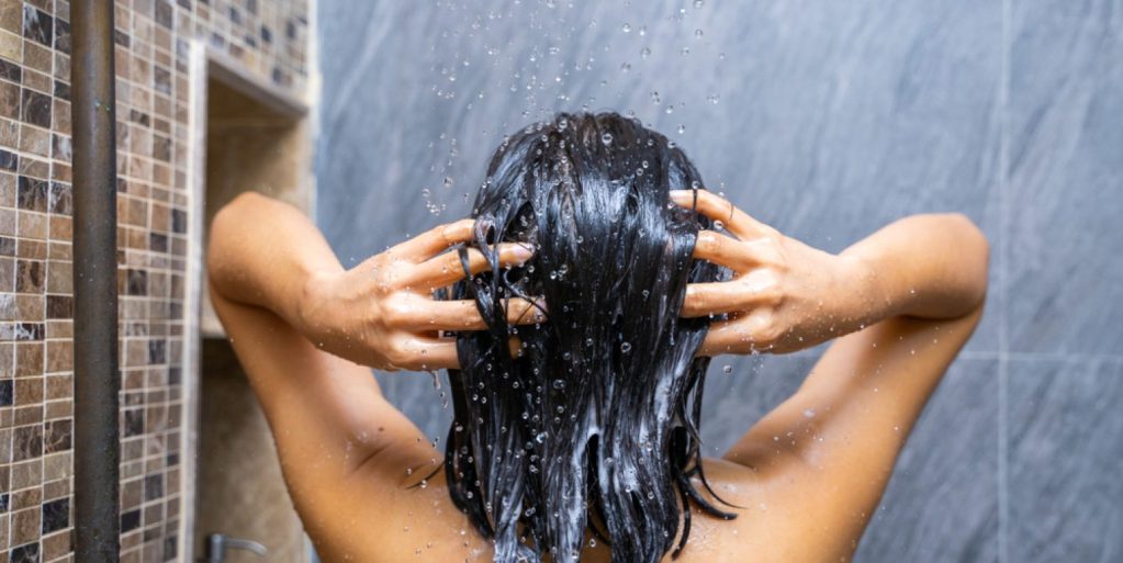Imagem vista de trás de mulher lavando o cabelo debaixo do chuveiro.