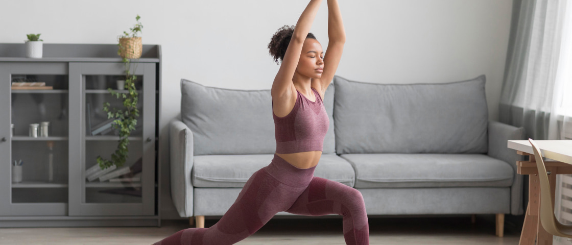 Imagem de uma mulher jovem, cabelos encaracolados presos com uma roupa roxa, fazendo exercícios de alongamento em um ambiente residencial com sala de estar, usando um tapete de yoga rosa, com uma parede branca, sofá cinza e janela com cortinas claras ao fundo.